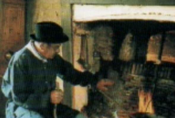 Un homme âgé en habit traditionnel attise le feu dans une grande cheminée quercinoise